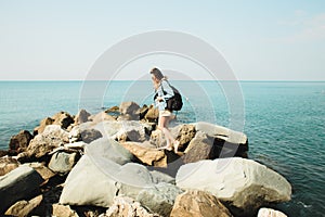 A young girl traveler with a backpack walks barefoot on large stones on the sea coast