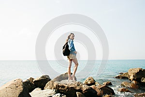 A young girl traveler with a backpack walks barefoot on large stones on the sea coast