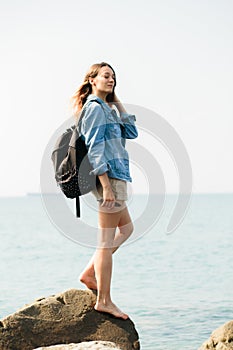 A young girl traveler with a backpack walks barefoot on large stones on the sea coast