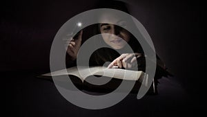 Young girl using flashlight in dark for reading book under the blanket