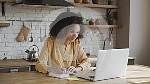 Young girl is using her laptop to study online, she is making notes while sitting on the kitchen at the table in light