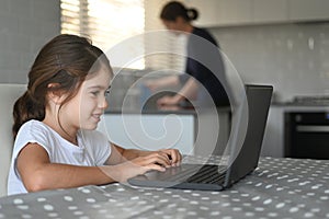 Young girl using a laptop at home while her mother cleaning the kitchen