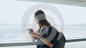 Young girl using smartphone near airport window. Happy European woman with backpack uses mobile app in terminal. 4K.