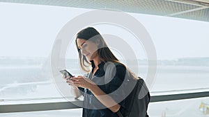 Young girl using smartphone near airport window. Happy European woman with backpack uses mobile app in terminal. 4K.