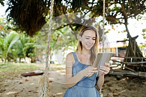 Young girl using tablet and riding swing on sand, wearing jeans sundress.