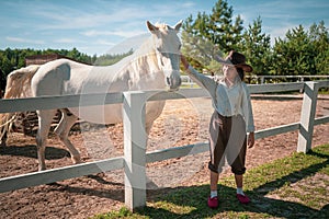Young girl in vintage clothes standing with her bright white horse in the paddock at sunny daytime. Tinted image