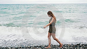 Young girl walking along the sea rocky coast. There are waves. The girl`s long hair is blown up by the wind.