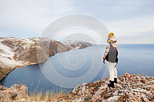 A young girl in warm clothes with a hiking backpack is standing at the cliff, admiring nature.