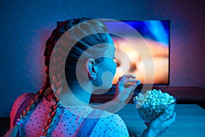 A young girl watching movies and eating popcorn with a bowl on the background of the TV. The color bright lighting, blue and red.