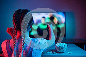 A young girl watching movies with a remote control with a bowl of popcorn on the background of the TV. A bright color of light,