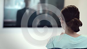 young girl watching tv and eating chips at home, brunette girl watching her favorite movie, back view