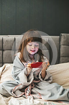 A young girl wearing a bathrobe sitting on a bed, engrossed in digital content with her smartphone in hands. Concept: kids and photo