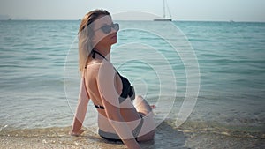 Young girl is wearing black bikini is relaxing on a beach in sunny day, back view, sitting in water