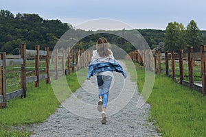 Young girl wearing a blue jeans and jeans jacket running outdoor. Girl running along the road in the stable.