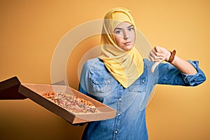 Young girl wearing muslim hijab holding box with Italian pizza over isolated yellow background with angry face, negative sign