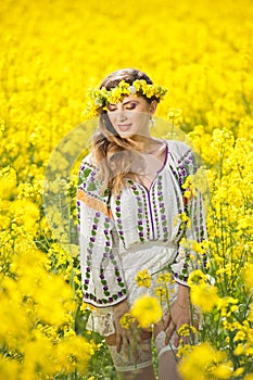 Young girl wearing Romanian traditional blouse posing in canola field, outdoor shot. Portrait of beautiful blonde with wreath