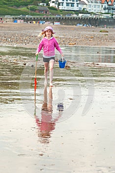 A young girl wears a pink floppy hat and carries a bucket and spade across a British beach