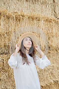 Young girl wears summer white dress near hay bale in field. Beautiful girl on farm land. Wheat yellow golden harvest in autumn