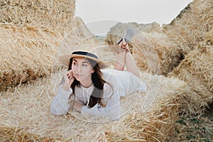 Young girl wears summer white dress near hay bale in field. Beautiful girl on farm land. Wheat yellow golden harvest in autumn