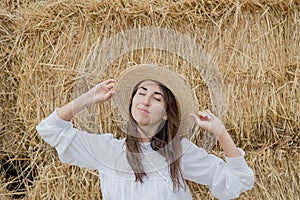 Young girl wears summer white dress near hay bale in field. Beautiful girl on farm land. Wheat yellow golden harvest in autumn