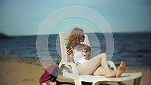 A young girl in a white bikini lies and sunbathes on a lounger on the sea sandy beach and uses smartphone
