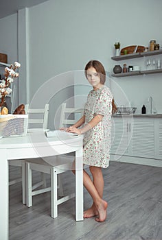 Young girl in white dress with long brown hair standing near table in white kitchen