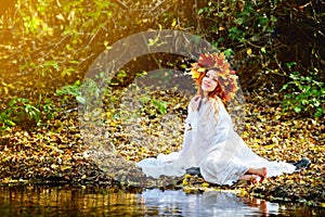 Young girl in white dress sitting on the river Bank in a wreath of yellow leaves in Sunny autumn day