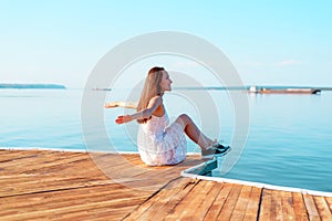 Young girl in white dress sitting on a wooden pier with open hands looking into the distance of the sea, freedom, clean air, dream