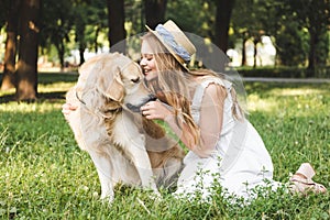 Young girl in white dress and straw hat feeding golden retriever while smiling and sitting on meadow with closed eyes