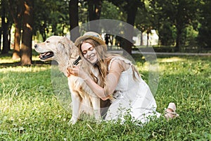 Young girl in white dress and straw hat hugging golden retriever while smiling and sitting on meadow with closed eyes