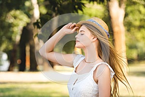 Young girl in white dress touching straw hat while standing in park with closed eyes