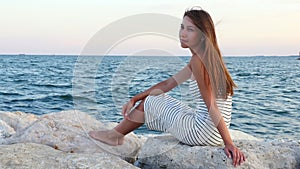 Young girl in white frock sits on rocks on the stone beach at the sea in sunset