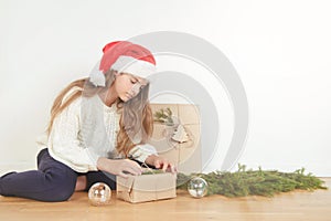 Young girl in white sweater and red santa hat wraps present with christmas tree branches close