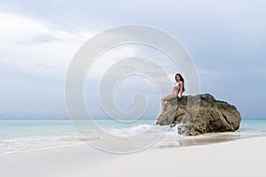 A young girl in a white swimsuit sits on a lonely stone on the beach. The waves hit the rocks