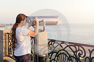 Young girl in white t-shirt looking through a coin operated binoculars on the sea shore. Woman look in touristic telescope on emba