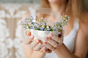 Young girl in a white tank holding a dotted cup with forget-me-not. Flowers in a mug. Hands with flowers.