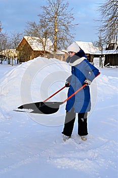 young girl in winter clothes cleans snow with shovel in countryside.