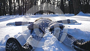 Young girl in winter clothes lying on snowy meadow and making angel. Happy woman playing in snow enjoying winter time