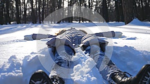 Young girl in winter clothes lying on snowy meadow and making angel. Happy woman playing in snow enjoying winter time