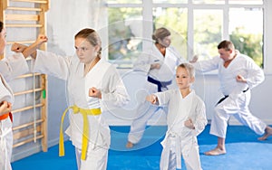 Young girl and woman in kimono and colored belt practicing karate punch block during group martial arts lesson in gym