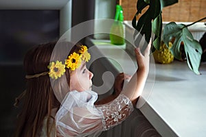 Young girl with a wreath of yellow flowers looking at a houseplant leaf on windowsill.