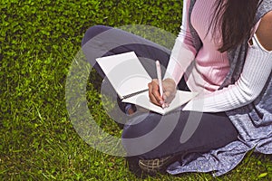 Young girl writes with pen in notebook sitting on green grass in park on meadow