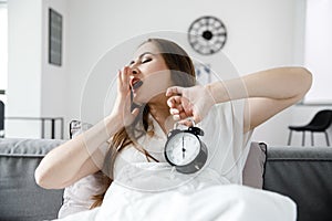 Young Girl Yawns with an Alarm Clock in her hands in a white, bright room in the morning