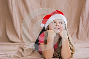 Little smiling girl in a red t-shirt with a santa claus hat lies on her stomach and smiles