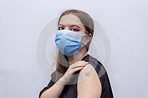 A young girl 18-20 years old in a medical mask points to a bandage with a vaccine against COVID-19, a coronavirus vaccination prog