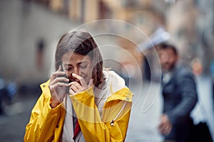 A young girl in a yellow raincoat is in a good mood while talking on the smartphone on the street on a rainy day. Walk, rain, city