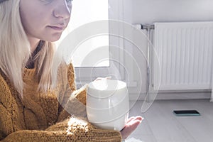 A young girl in a yellow sweater and hat is drinking hot tea, near a heater with a thermostat.