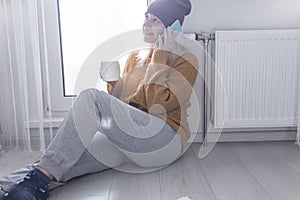 A young girl in a yellow sweater and hat is talking on the phone and drinking hot tea, near a heater with a thermostat