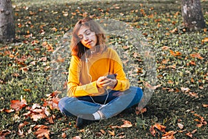 Young girl in yellow sweater listening to the music with earphones sitting on autumn grass