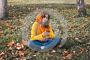 Young girl in yellow sweater listening to the music with earphones sitting on autumn grass